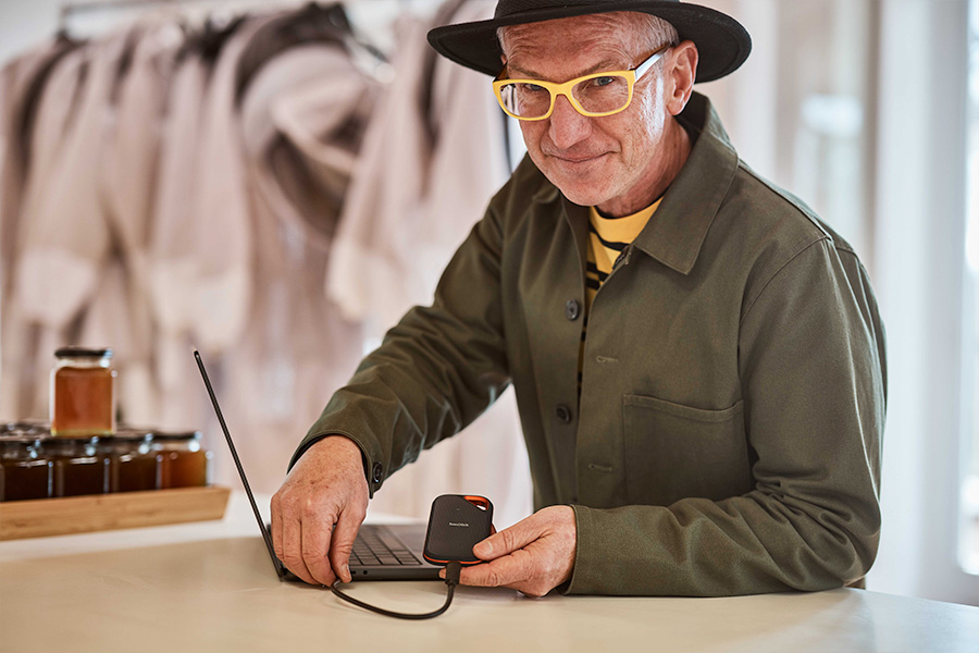 Hamish using a laptop connected to an external hard drive, with jars of honey and beekeeping suits visible in the softly focused background.