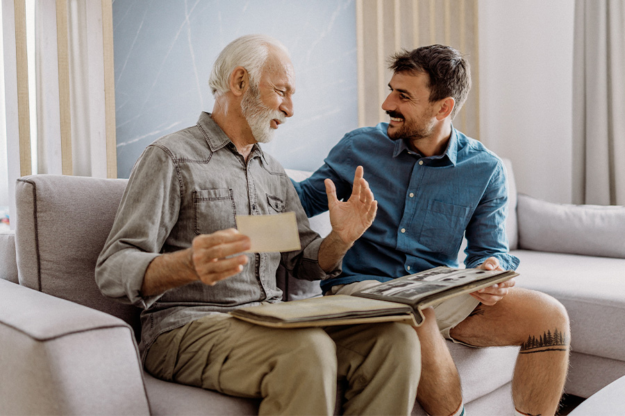 Two people sitting on a couch, engaging in conversation while looking at a photo album together. A modern living room setting is visible in the background.