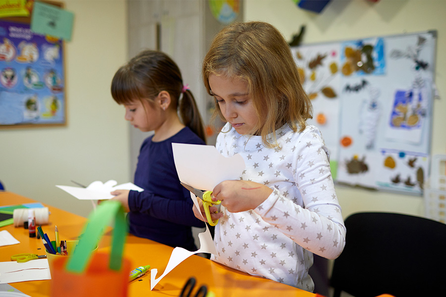 Two primary-school-aged children at an orange table in a classroom, cutting white paper with scissors, with pencils and craft supplies scattered nearby.