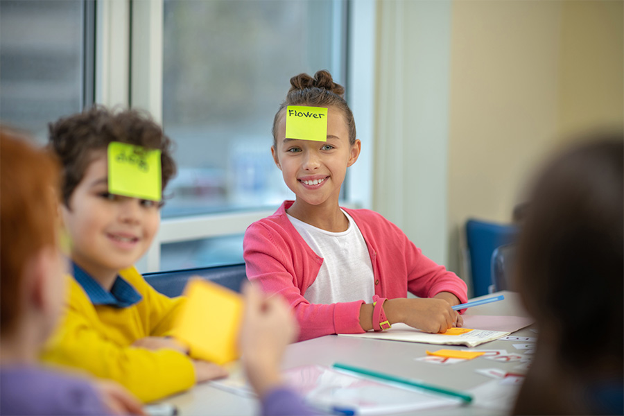 Primary-school-aged students seated at a classroom table near a window; a student writes in a notebook while classmates discuss an activity with sticky notes and letters.