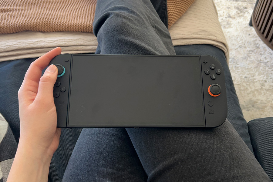 A close-up of a hand holding a Nintendo Switch 2 gaming console with a blank screen, resting on stretched-out legs on a sofa.