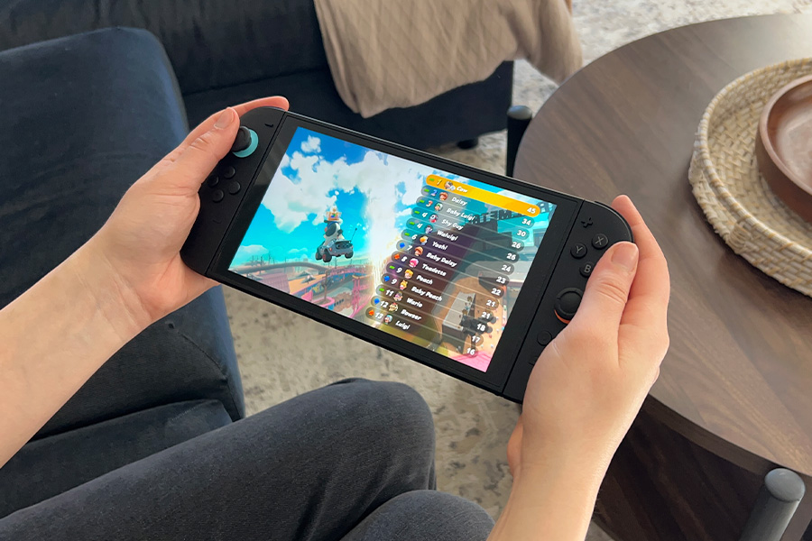  Hands holding a Nintendo Switch 2 gaming console displaying a Mario Kart game leaderboard, with a sofa, round wooden table and woven tray visible in the background.