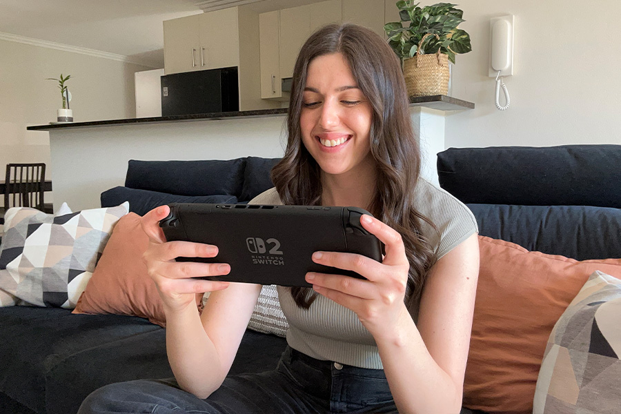 Marissa playing on a Nintendo Switch 2 console while seated on a sofa, with cushions, a kitchen counter and a potted plant visible in the background.