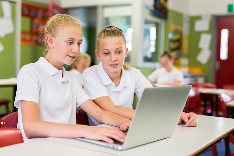 Two high-school-aged students working together on a laptop in a classroom with colourful decorations and desks in the background.