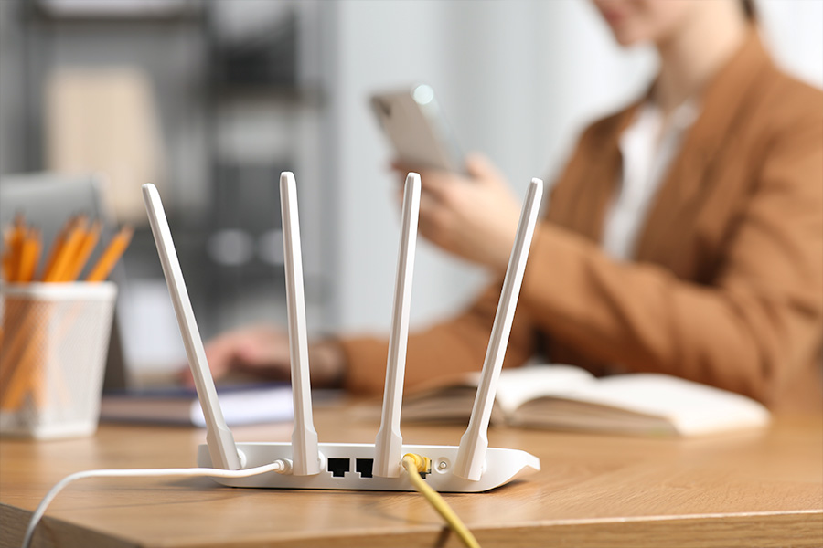  A white Wi-Fi router with multiple antennas on a desk, connected by cables, with a blurred person in the background sitting at the desk using a smartphone.