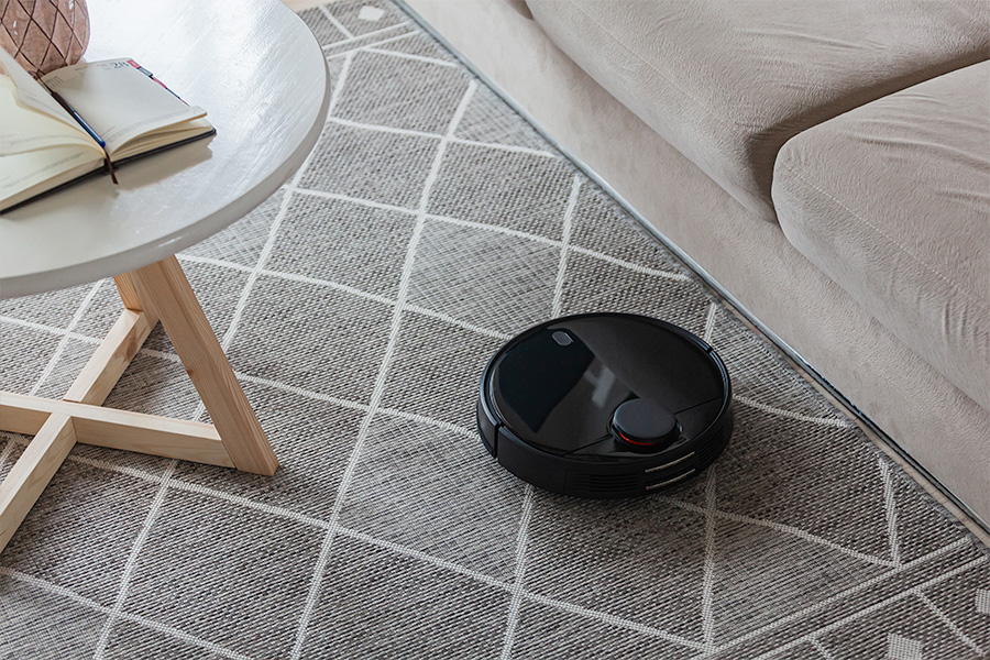 A black robotic vacuum cleaner operating on a patterned carpet near a beige sofa and a wooden coffee table with an open notebook placed on top.