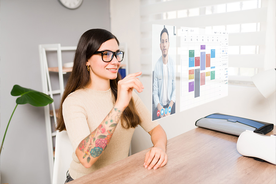 A person wearing smart glasses interacting with a futuristic virtual screen displaying a video call and a digital calendar in a modern office setting with a desk and indoor plant.
