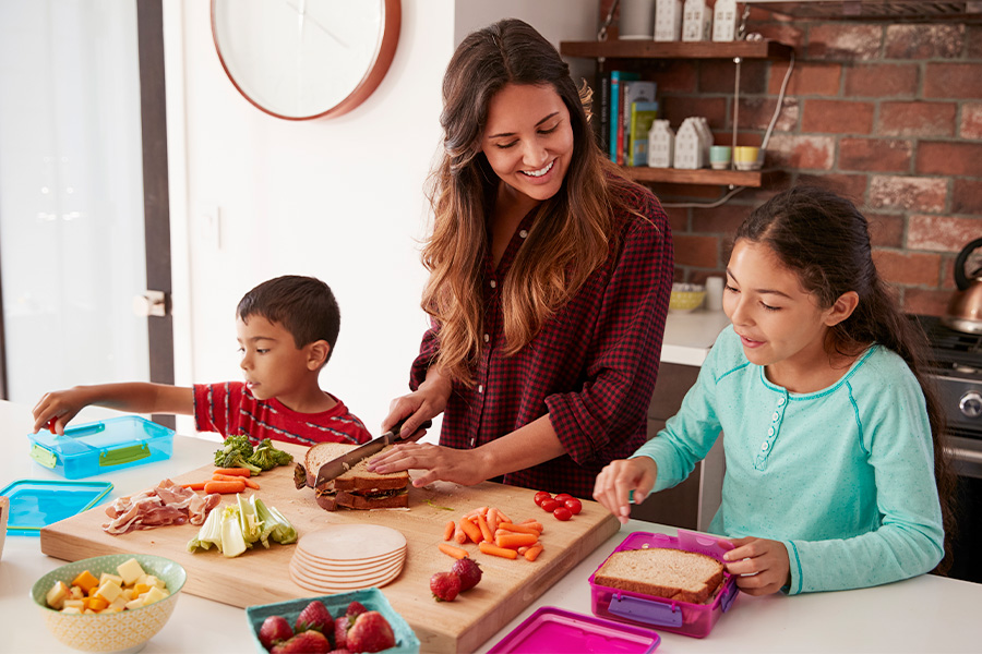 Two children and an adult packing lunch boxes in a bright kitchen setting, illustrating the benefit of kids' participation in meal preparation.