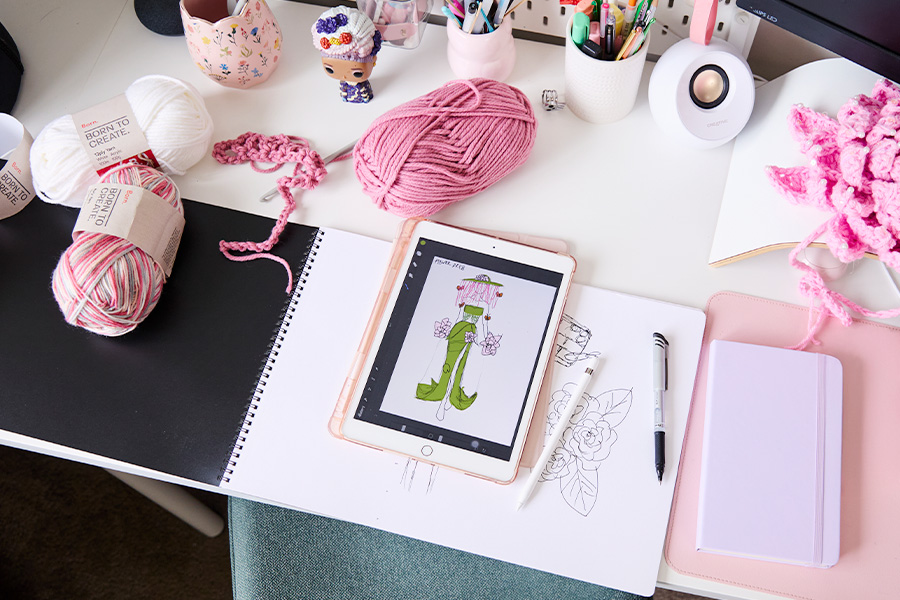 A tablet showing a digital fashion sketch on a white desk, surrounded by a sketchpad, pink yarn, crocheted pieces, pens and notebooks.