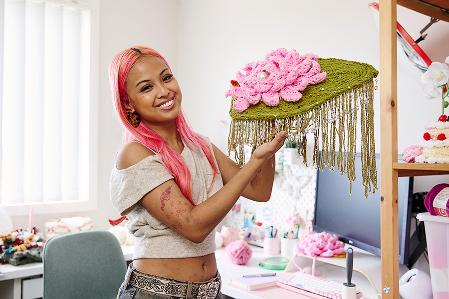 Katelyn holding a green crochet headpiece adorned with a large pink flower and beaded fringe in her home craft studio.