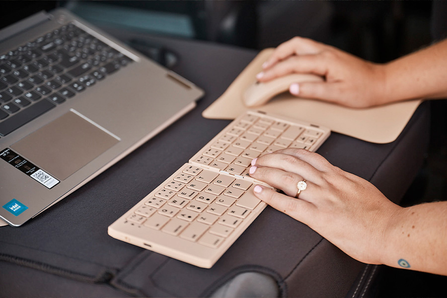Close-up of hands using a beige keyboard and mouse on a folded-down seat, with a laptop on a laptop stand nearby.