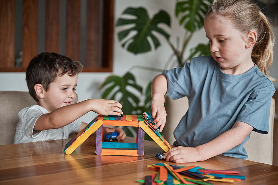 Two children playing on a bridge made from colourful craft sticks at a table, racing small toy cars up the ramps in a cosy living room setting.