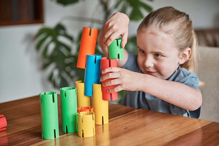 A child carefully stacking green, blue, yellow, orange and red slotted cardboard rolls to create a tower on a wooden table in a living room setting.
