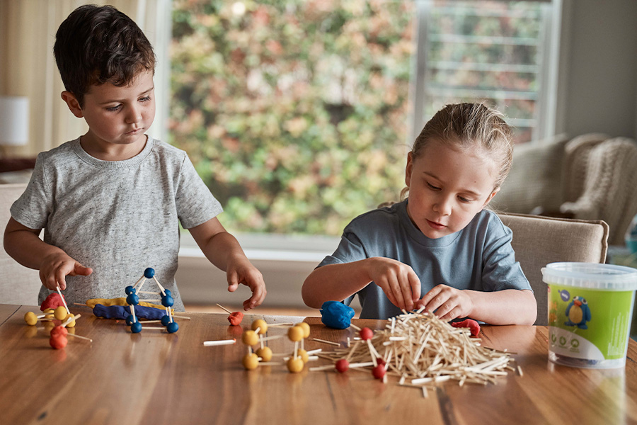 Two children building with playdough and craft matchsticks at a table, rolling balls and connecting them into shapes beside a pile of sticks in a bright living room setting.