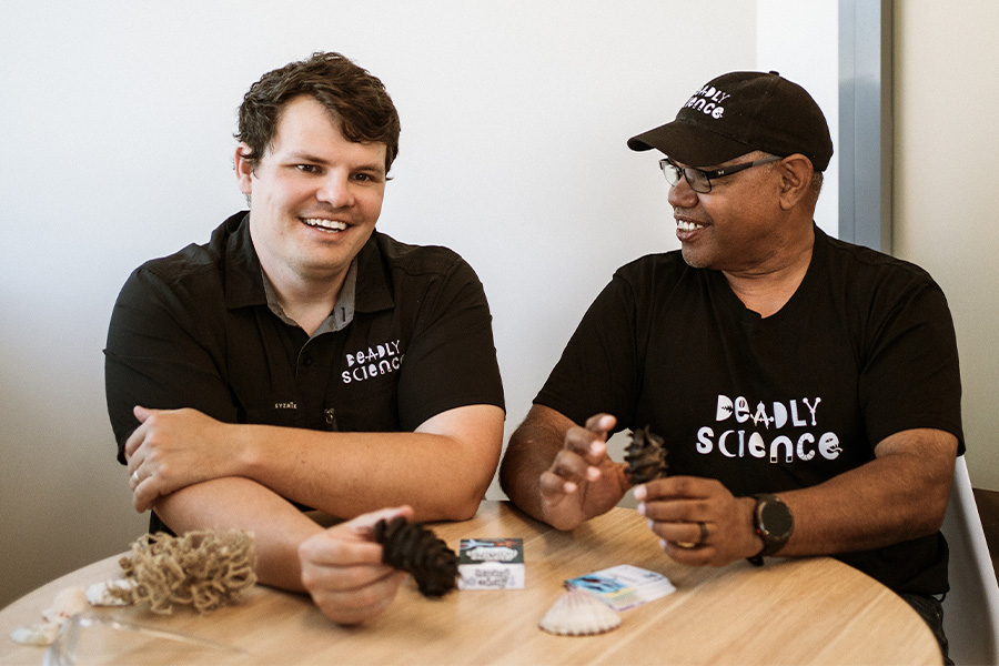 Corey and Vince sit at a table examining sea specimens, including shells, moss, coral and shark eggs, with cards and notes nearby.