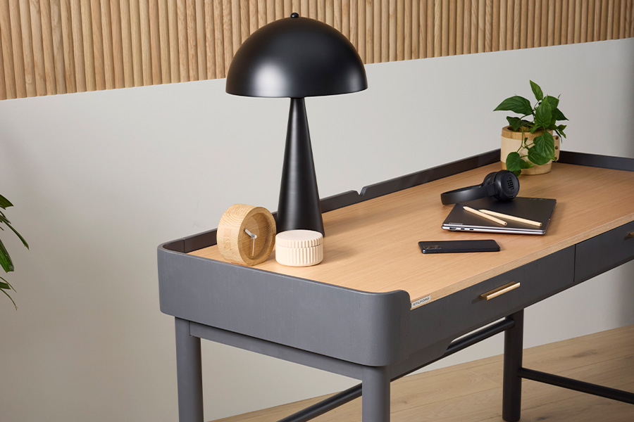 Close-up of a modern desk with light timber top and grey frame, black dome lamp, potted plant, headphones, laptop, notebook, pencils and phone.