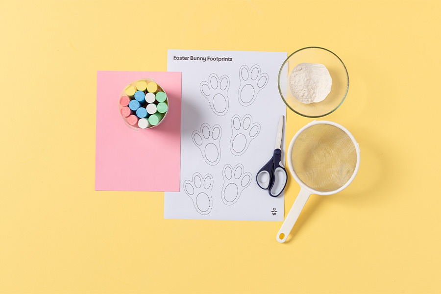An assortment of supplies to help with creating bunny footprints, arranged on a yellow background. Products include bunny footprints printed on paper, chalk, scissors, flour and a sifter.