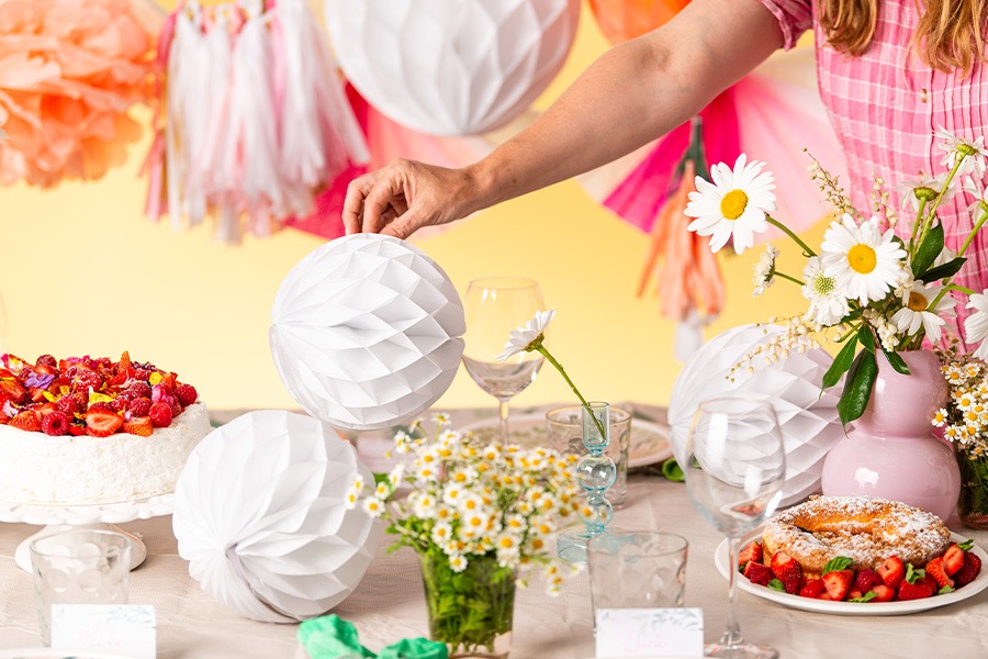 A person setting an Easter holidays tablescape with white honeycomb paper decorations, fresh daisies and two delicious-looking cakes topped with berries.
