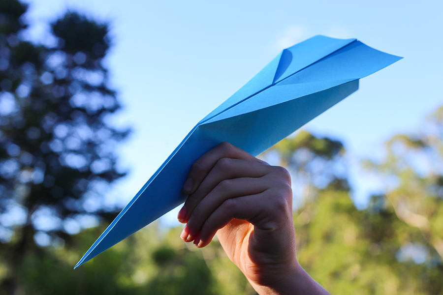 A hand holding a basic dart paper plane made from blue paper outdoors against a clear sky, with blurred trees in the background.