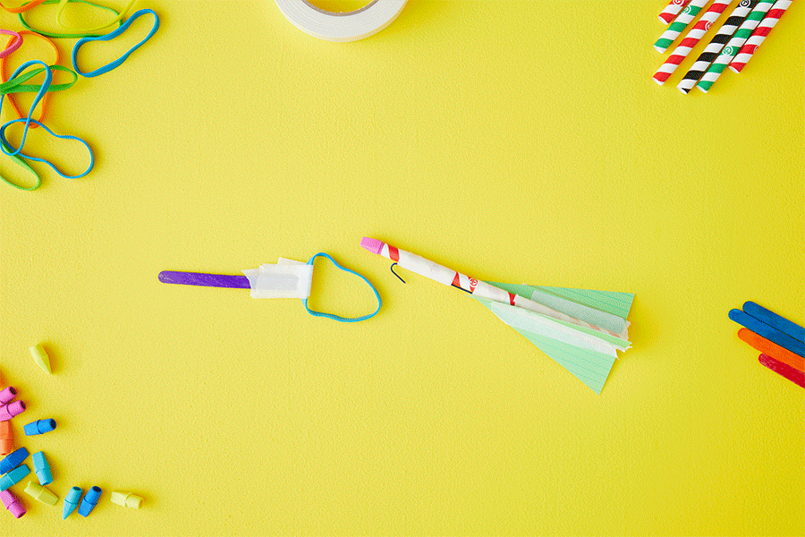  A series of images showing a slingshot straw rocket STEM activity: a rocket made of a straw, eraser, paperclip and cardboard is launched across a yellow background using a slingshot made of a rubber band taped to a craft stick.