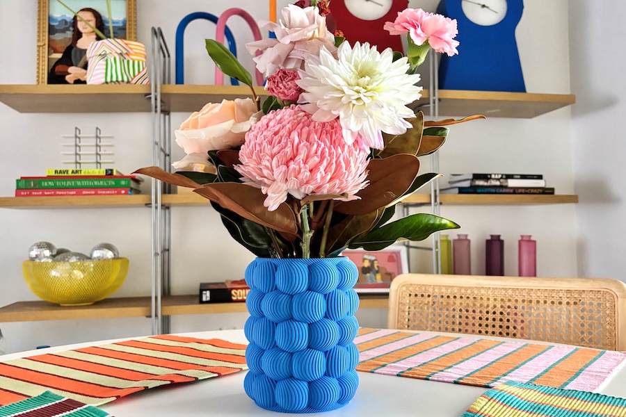 A blue printed 3D vase full of pink and white flowers and green leaves sitting on a table with a bookshelf in the background covered in colourful home decor items.