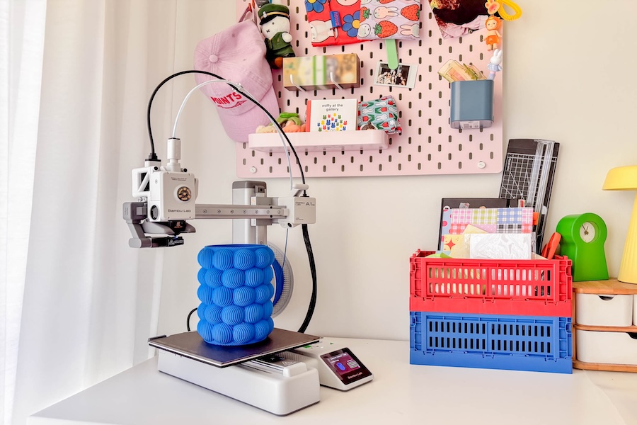A blue printed 3D vase standing on the platform of the Bambu Lab A1, standing on a white desk with stationery items to the right and a pink pinboard covered in home decor items.