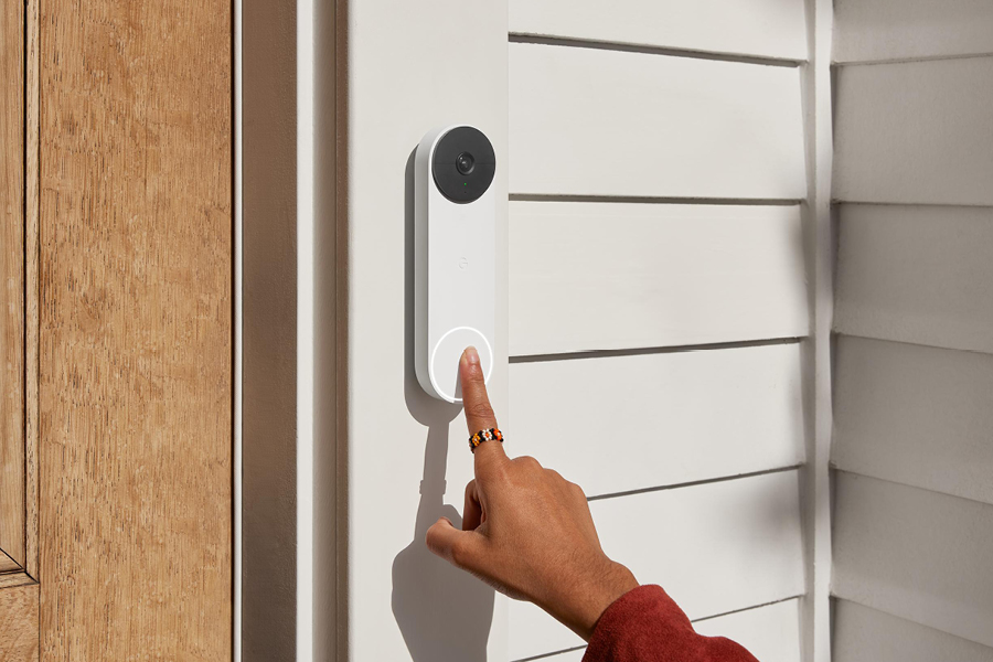 A finger presses a Google Nest smart doorbell mounted to a white door frame of a white wooden house with a light brown wooden door.