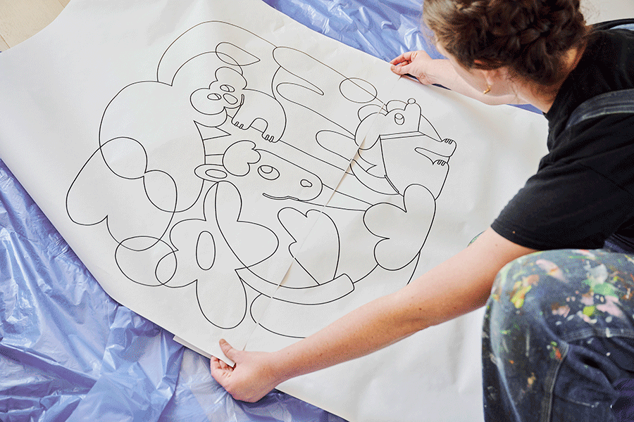 A series of images showing Lydia sketching, holding, taping and positioning a large cartoon wall mural stencil, from rough draft to clean outline.