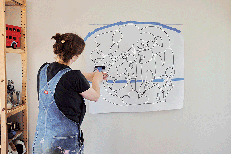 A series of images showing Lydia rubbing over a taped stencil to transfer a cartoon mural outline onto a wall, revealing red guidelines, then tracing the lines by hand.