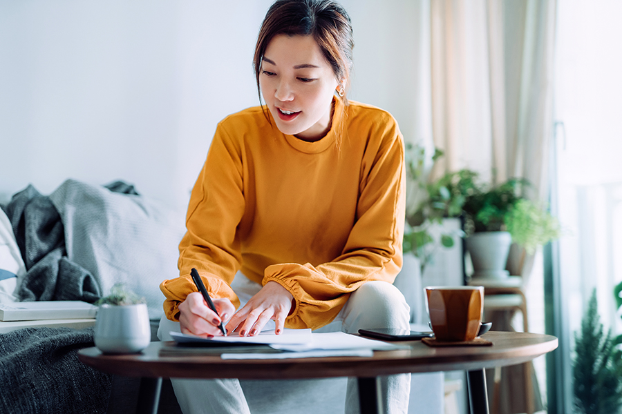 A woman in a yellow top leans over a coffee table to write on sheets of paper. A brown coffee cup is to her side.