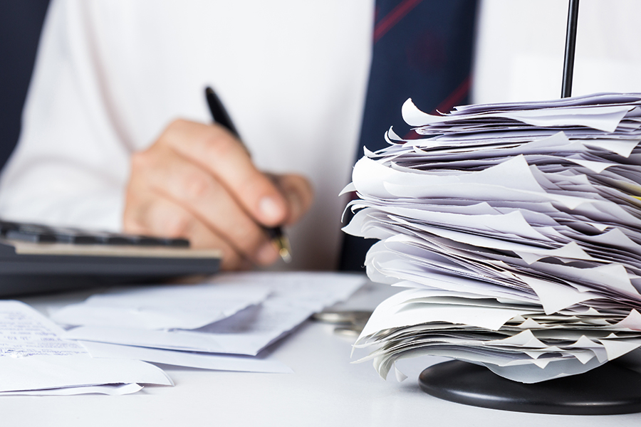 A receipt folder stacked with invoices is in the foreground, while a figure in a shirt and tie holds a pen to write on paper in the background.