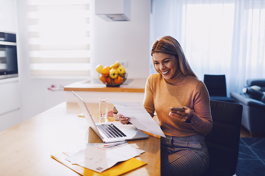 A smiling woman sits at a kitchen table in front of an open laptop and assorted bills, holding an invoice and a phone in her hands.
