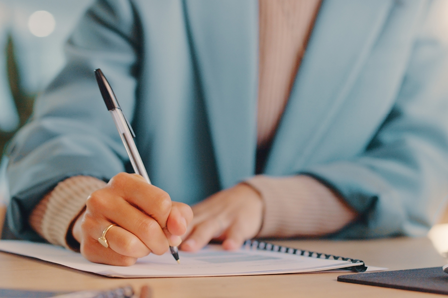 A close-up view of a person’s hand writing in a booklet using a pen. The person is sitting at a desk and wearing a light blue coat.