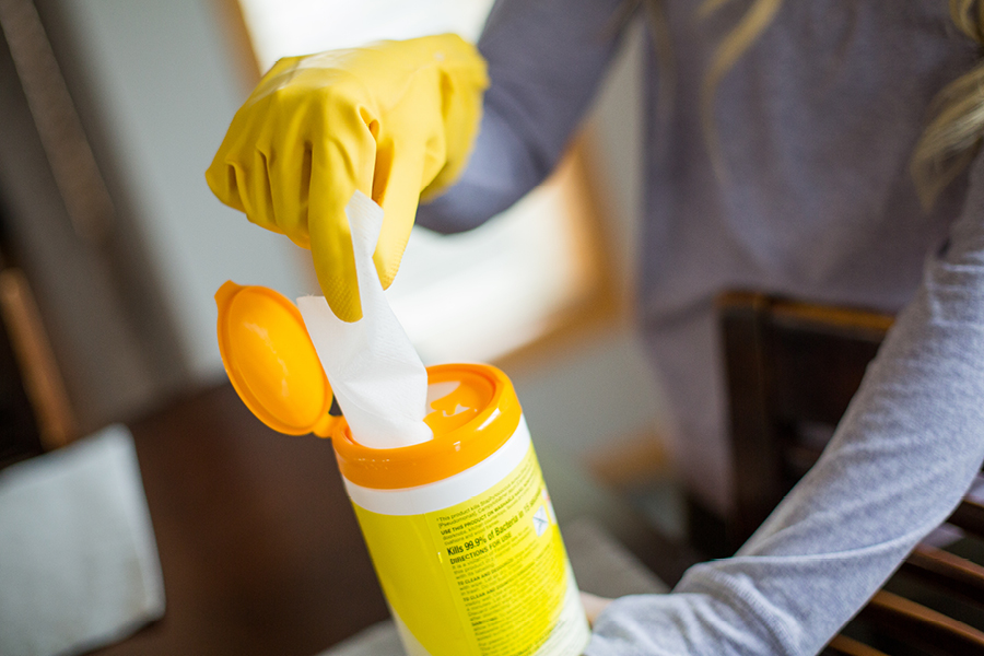 A person wearing a yellow rubber glove pulls a disinfectant wipe from an orange-lidded canister.