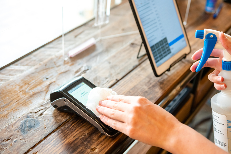A hand cleaning an EFTPOS terminal on a wooden counter using a paper towel, with the other hand holding a spray bottle, and a point‑of‑sale tablet also on the counter.