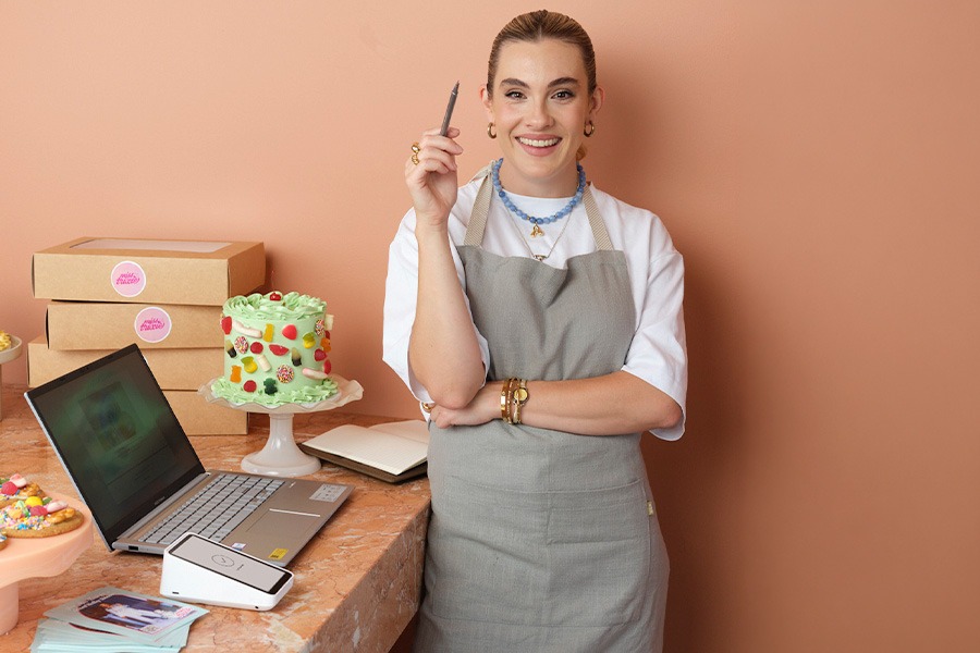 Alice standing, holding a pen next to a marble counter with laptop, card reader, notebook, boxed cakes and a green fruit‑decorated cake on a stand, against a peach wall.