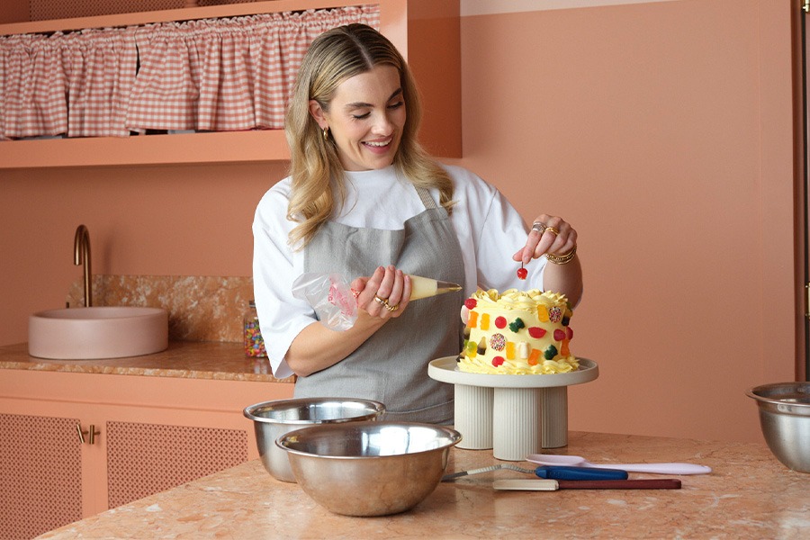 Alice piping yellow icing onto a fruit-decorated cake on a stand in a peach-coloured kitchen, with mixing bowls and spatulas on the marble bench.