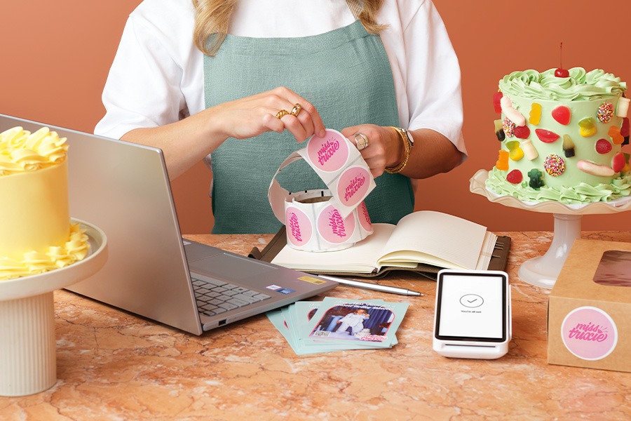 A close-up of Alice’s hands holding a roll of pink round stickers that say ‘Miss Trixie’ at a marble counter, with laptop, open notebook, card reader and green decorated cake nearby.