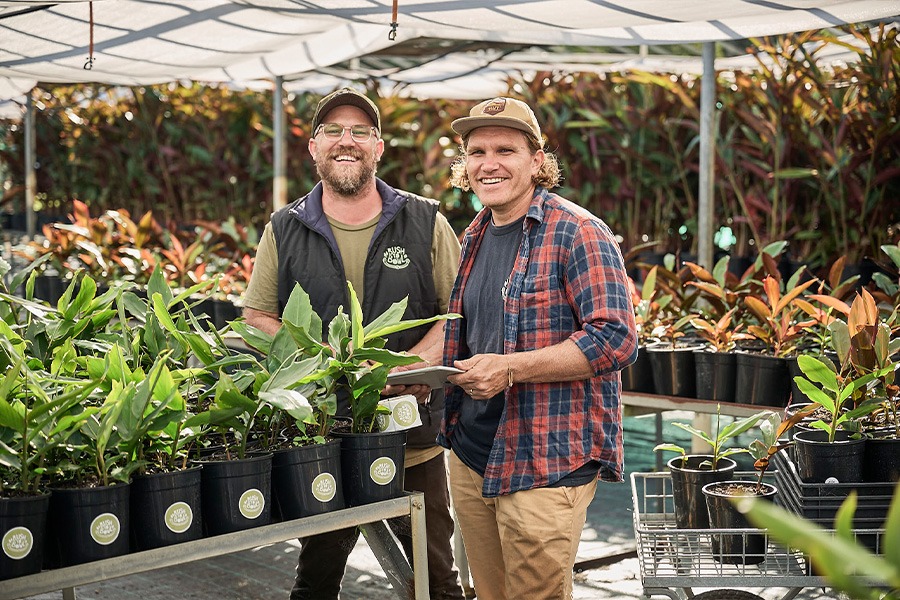 Adam and Clarence standing in their nursery surrounded by rows of potted plants under shade cloth; Clarence holds a tablet.