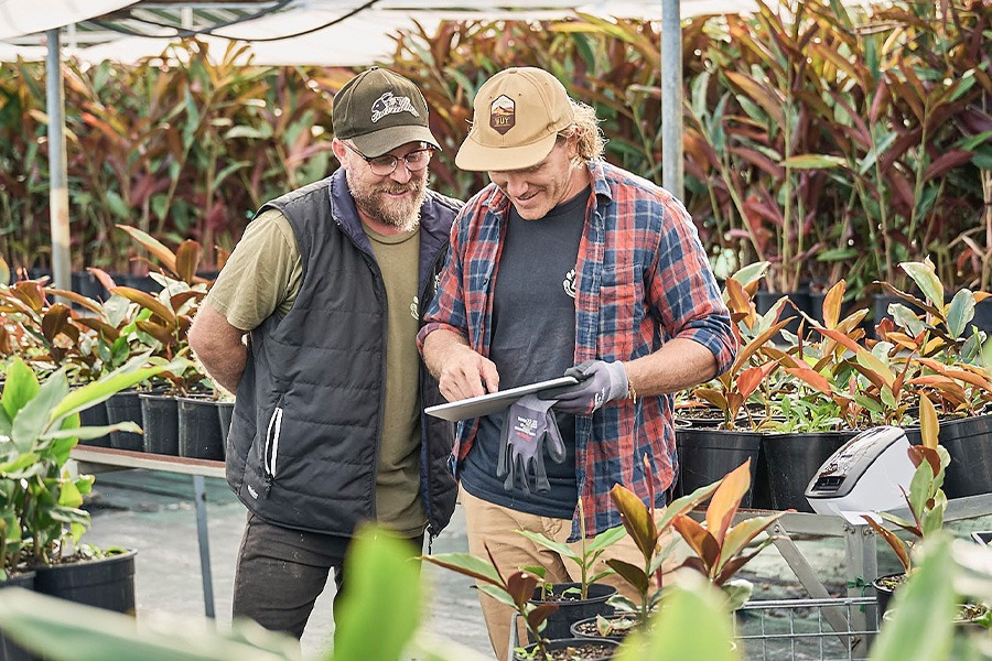 Adam and Clarence review a tablet among rows of potted plants under shade cloth; a label printer sits on a nearby bench.