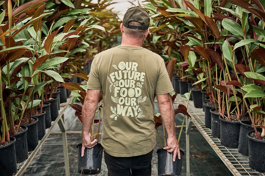 Adam from behind in an olive T-shirt reading ‘OUR FUTURE OUR FOOD OUR WAY’ walking between rows of potted plants, holding two black pots.