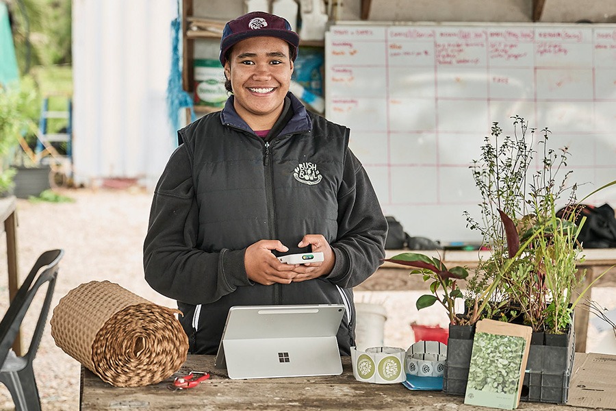 A Bush to Bowl employee standing behind a table holding a credit card reader, with a 2-in-1 laptop, label rolls, rolled paper and trays of native seedlings.
