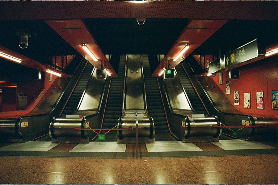 A 35mm film photo of an empty red‑walled underground Hong Kong station with a bank of escalators, some roped off with red barriers; fluorescent lights and signage overhead.