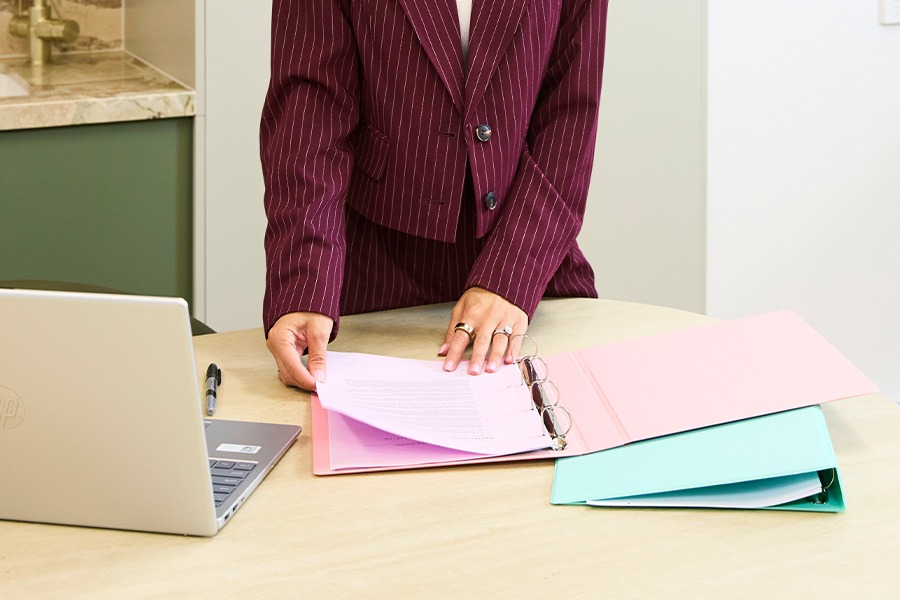 A person flipping through documents in a pink ring binder on a meeting table, with a laptop, pen and a teal folder nearby.