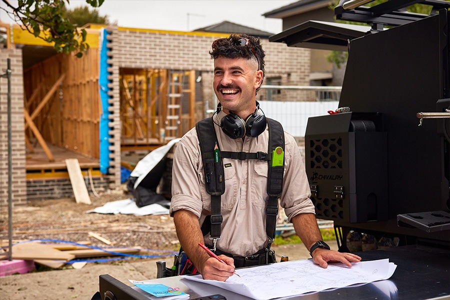 Tristan using a carpenter’s pencil to mark site plans at a ute‑mounted workstation on a house construction site.