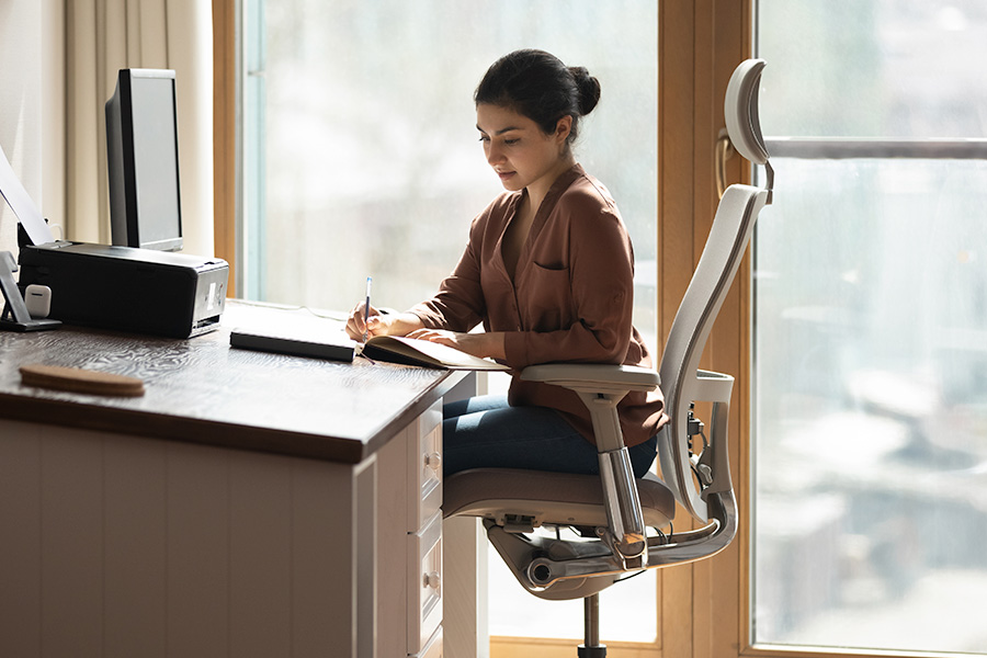 A person sits in an ergonomic office chair at a home office desk, writing in a notebook beside a computer and printer, with daylight from a large window behind.
