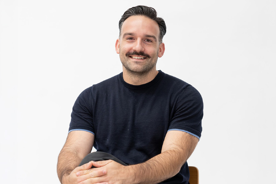 Julian Mauro seated with hands clasped, smiling at camera against white background in a studio setting.
