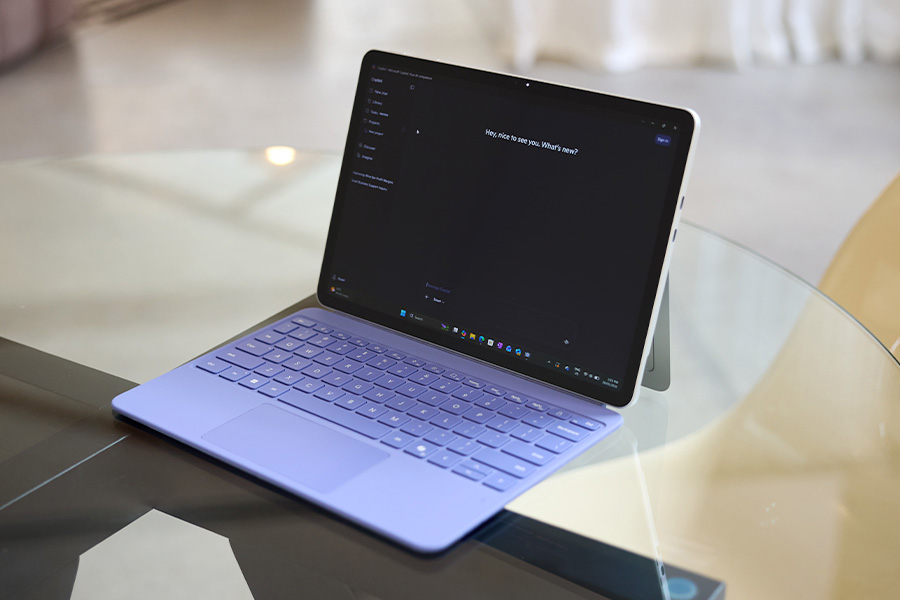A Microsoft Surface Pro in laptop mode on a glass table with a lavender keyboard; screen shows Copilot on a dark theme, supported by a rear kickstand.