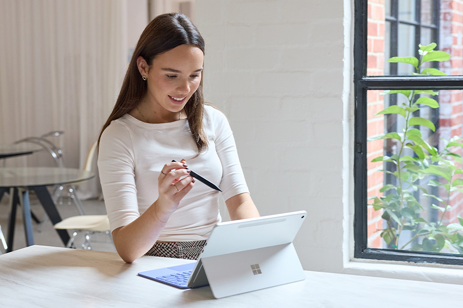 Kelsie sitting by a window holding a stylus above a Microsoft Surface Pro on a table; the tablet rests on its kickstand with a lavender keyboard, in a bright studio workspace.