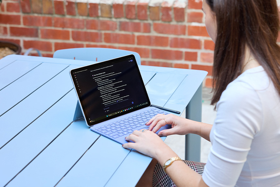Kelsie typing on a Microsoft Surface Pro with a lavender keyboard on a light blue outdoor table; tablet on its kickstand with a stylus beside it, brick wall in the background.